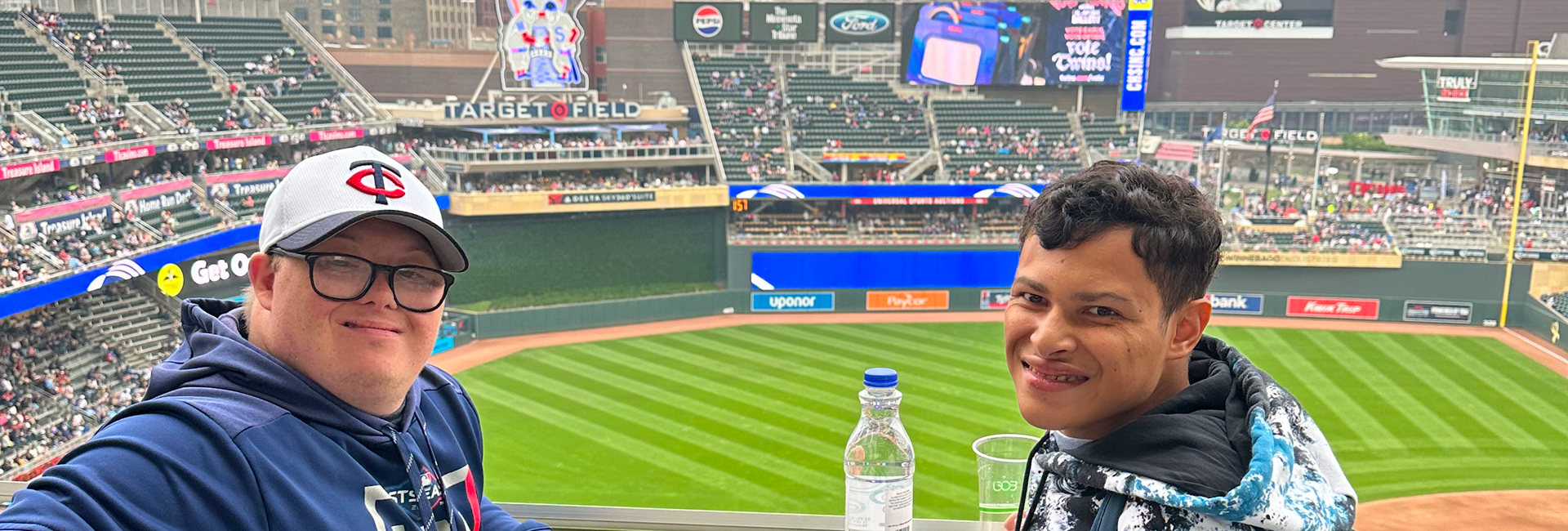 A person smiling at a baseball stadium with a water bottle in front.
